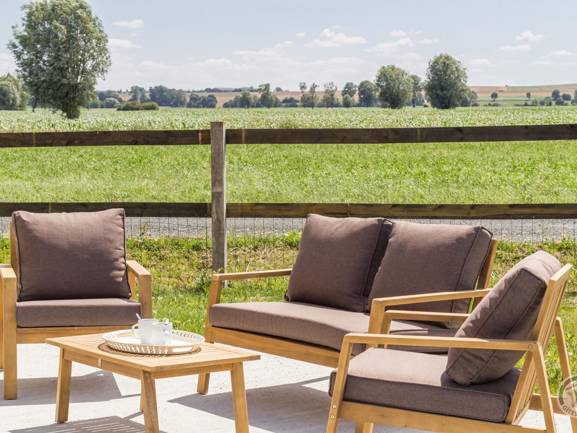 An outdoor seating area with a wooden table and two sofas, a wooden fence, and a view of a green field.