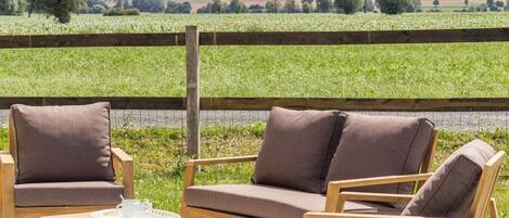 An outdoor seating area with a wooden table and two sofas, a wooden fence, and a view of a green field.