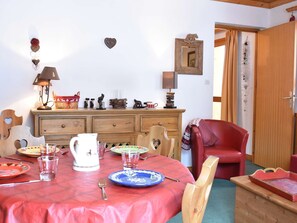 Un coin repas avec une table en bois dressée pour deux personnes, une chaise rouge et un meuble en bois orné d’objets décoratifs.