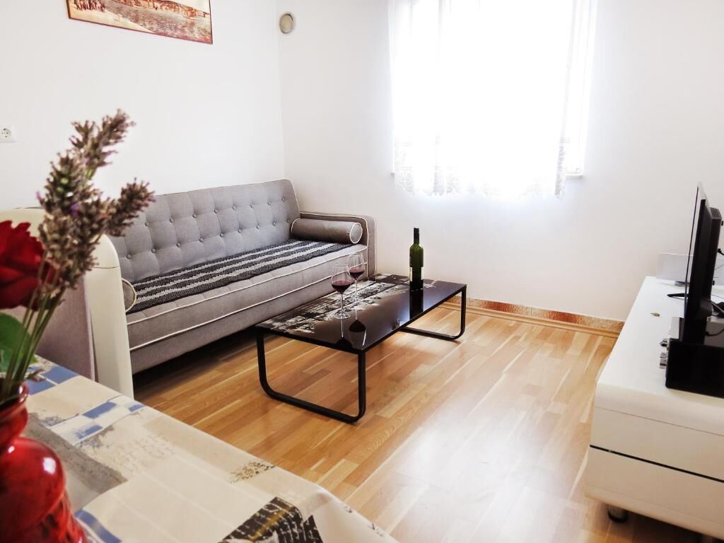 A living room with a grey sofa, a black coffee table with a wine glass, a red vase with flowers, and a white cabinet with a TV.