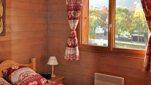 A wooden cabin room with a bed, a nightstand, a lamp, a window with curtains, and a heater. - 3-room apartment with large balcony, parking (La Chapelle-d'Abondance)