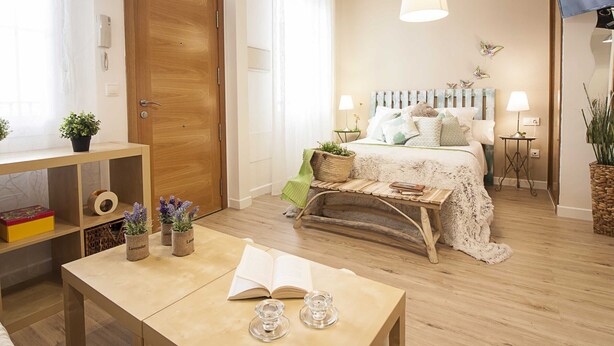 Un dormitorio acogedor con una puerta de madera, una mesita de noche con un libro y cristalería, una cama con cabecera blanca y azul, y una lámpara colgante.