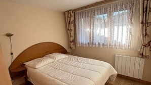 A bedroom with a wooden headboard, a white bedspread, a window with patterned curtains, and a radiator. - Apartment with Sunny Terrace in La Chapelle-d'Abondance (La Chapelle-d'Abondance)