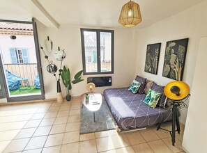 A living room with a sofa, a small table, a potted plant, a hanging light fixture, and a window with a view of the outside.