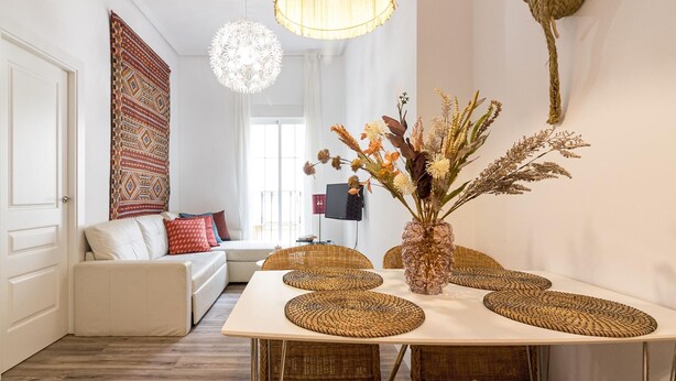 A modern living room with a white sofa, a dining table with woven placemats, and a decorative vase with dried plants. - Bright, Newly Renovated 2BR Apt in Historic Center Near Calle Larios (Málaga)
