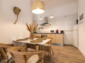 A modern kitchen with a dining area, featuring a white table, wicker chairs, and a decorative vase with dried flowers.