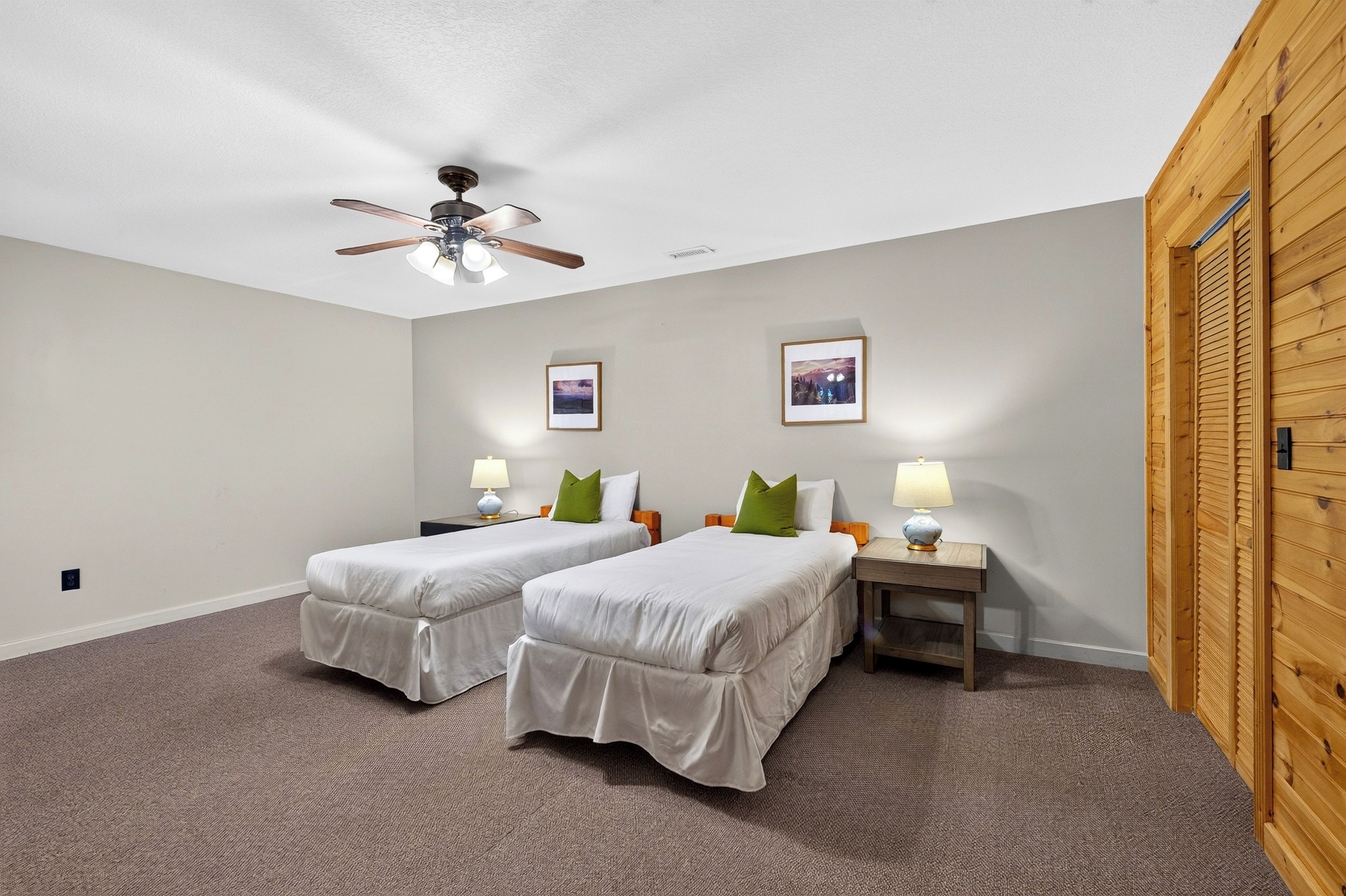 Habitación de hotel con dos camas, un ventilador de techo, dos mesitas de noche con lámparas y dos cuadros en la pared.