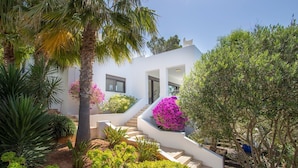Une maison moderne à l’extérieur blanc, avec un escalier menant à l’entrée, et diverses plantes et arbustes qui ornent la propriété.