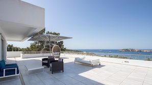 Une terrasse dotée d’un fauteuil suspendu, de deux canapés et d’une table placée sous un grand parasol. La terrasse offre une vue sur la côte, avec des bateaux et une île au loin.
