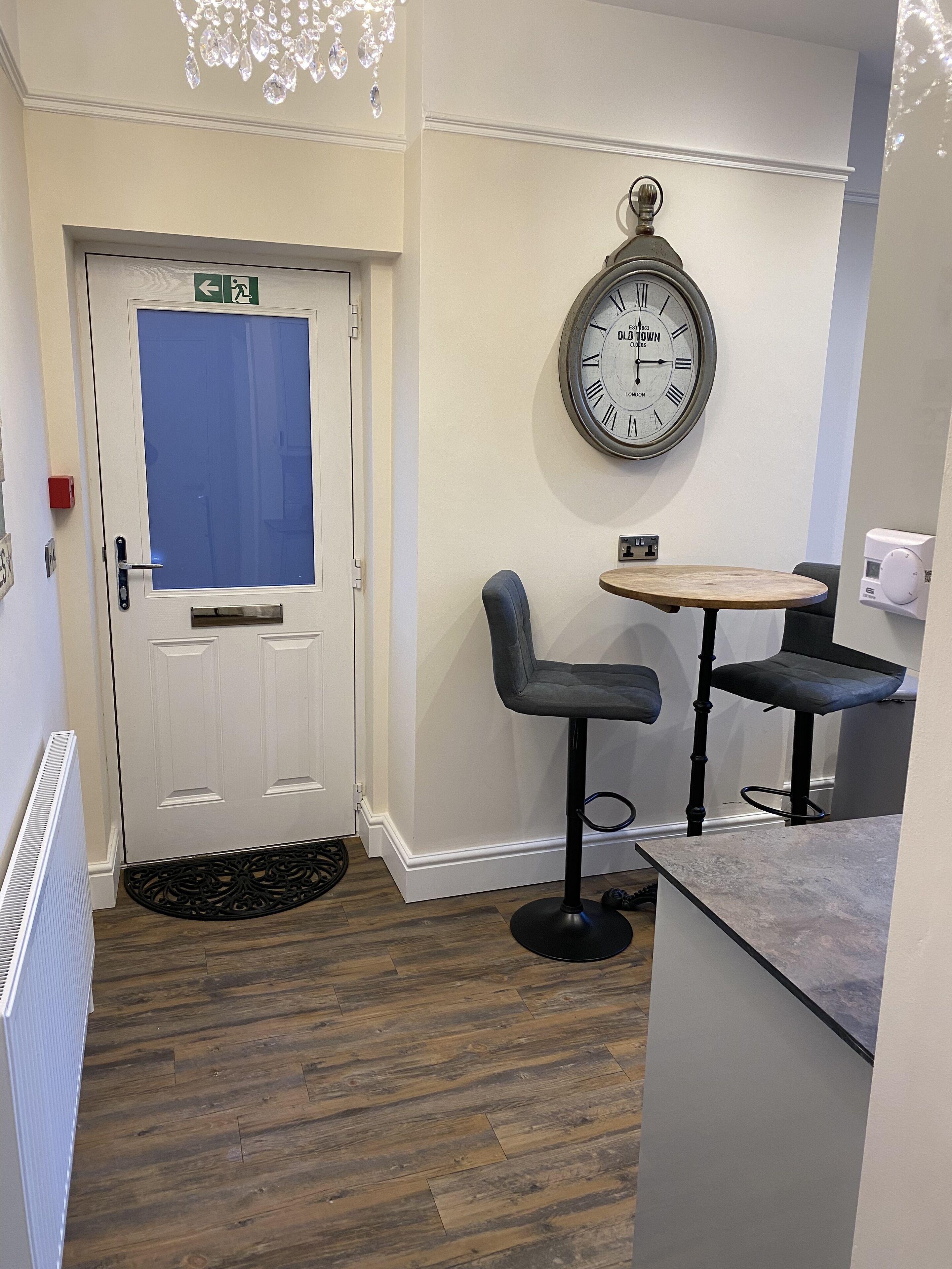 A room with a door, a wall clock, two bar stools, a small table, and a radiator.