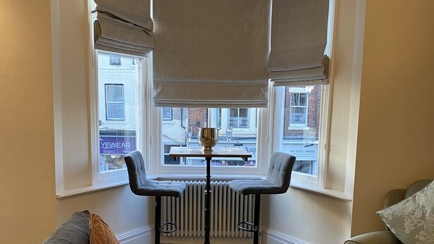 Ein Eckzimmer mit Erkerfenster, Radiator, einer Chandelierleuchtre, einem Barhocker, einer Couch und einem Couchtisch.