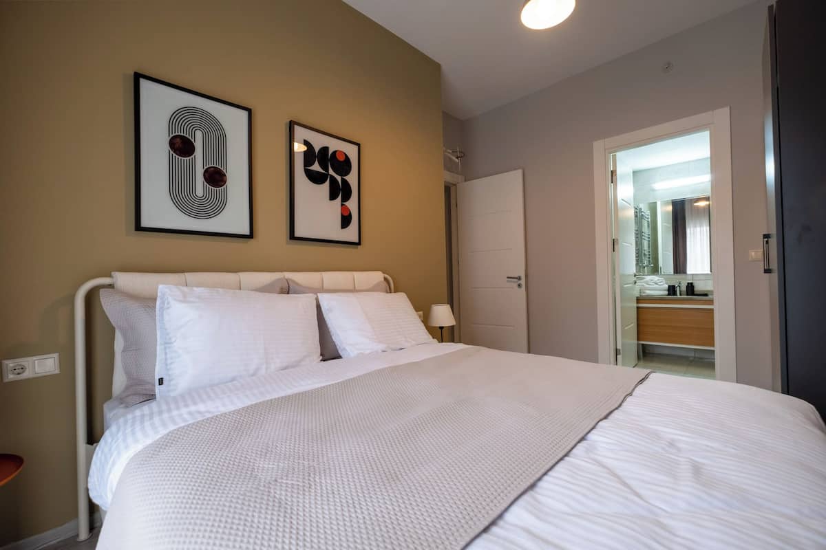 A neatly made bed with white bedding, a bedside table, and two framed artworks on the wall.