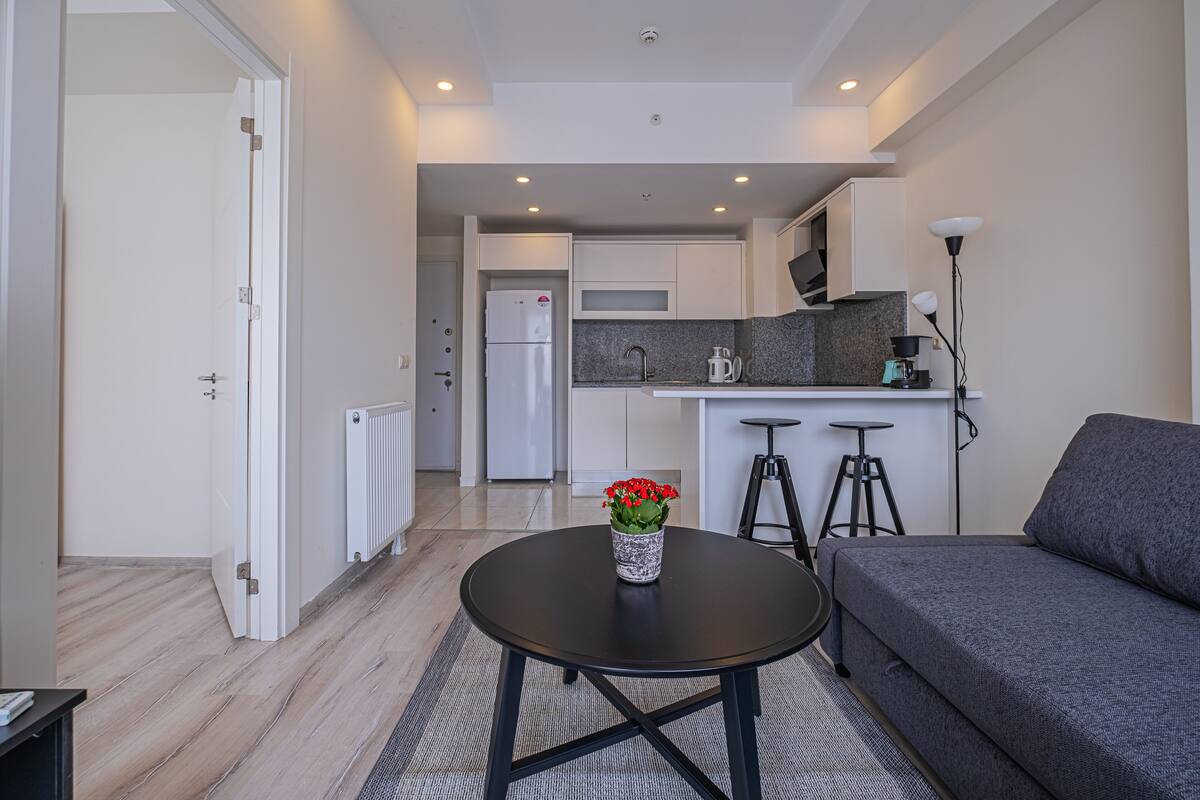 A modern living room with a sofa and a coffee table, a kitchen area with a refrigerator, microwave, and bar stools, and a door leading to another room.