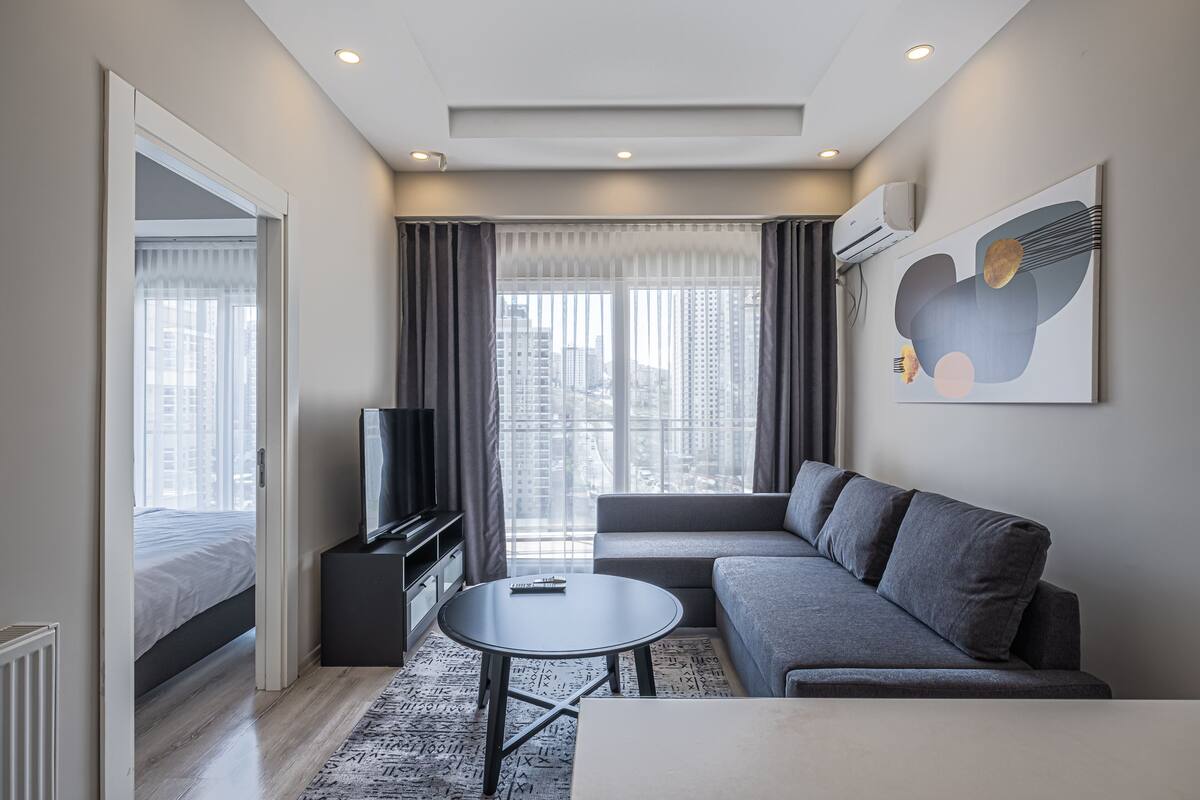 A modern hotel room with a large window, a sofa, a TV, and a small table.