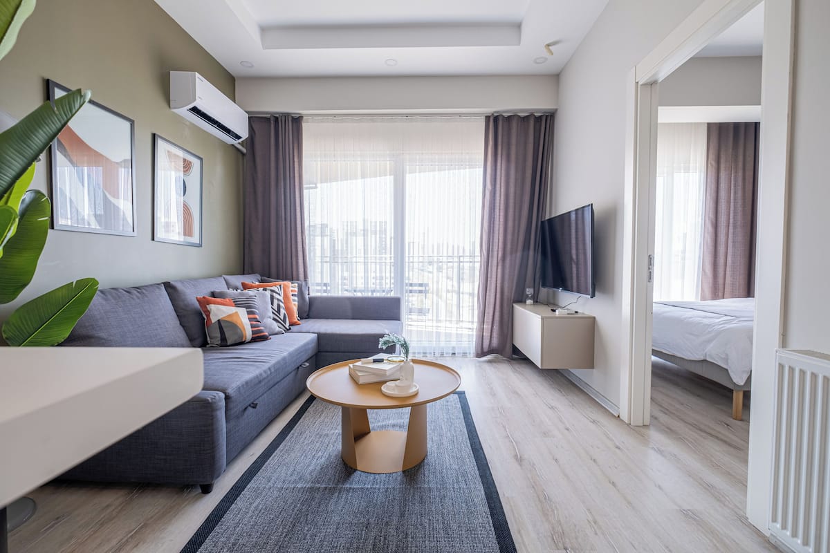 A modern hotel room with a grey sofa, a round coffee table, a flat-screen TV, and a balcony with curtains.
