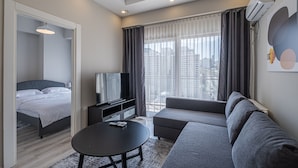 Habitación de hotel moderna con cama, televisor, sofá y un amplio ventanal con cortinas.