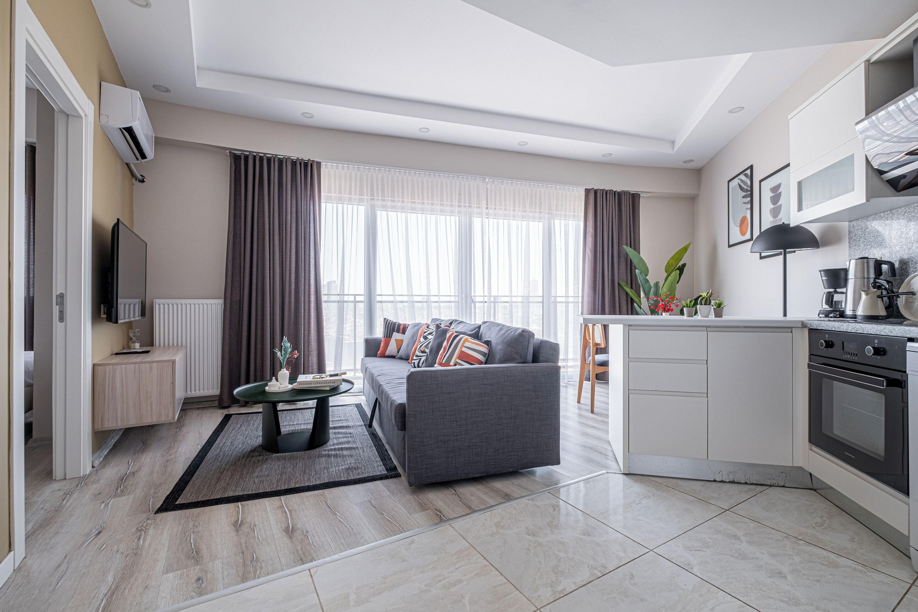 Ein modernes Wohnzimmer mit grauem Sofa, einem Couchtisch und einem Fernseher. Eine Küchenzeile mit Herd und Schränken ist ebenfalls vorhanden.