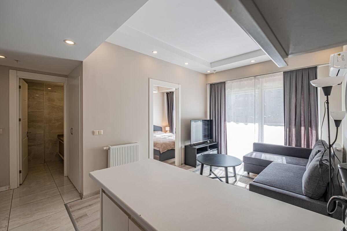 A modern hotel room with a kitchenette, a sofa, a TV, and a large window with curtains.