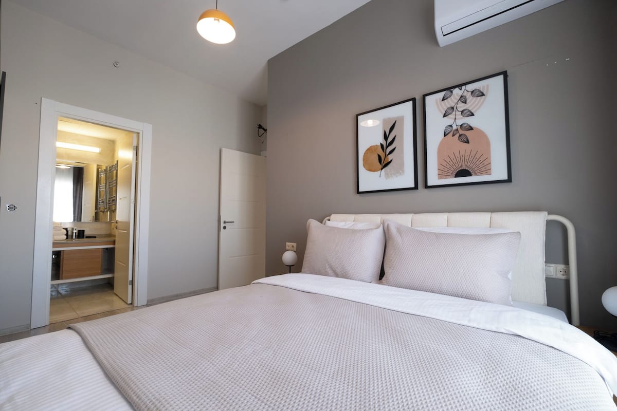 A bedroom with a bed, a bedside table, a nightstand, a mirror, and a door leading to a bathroom.