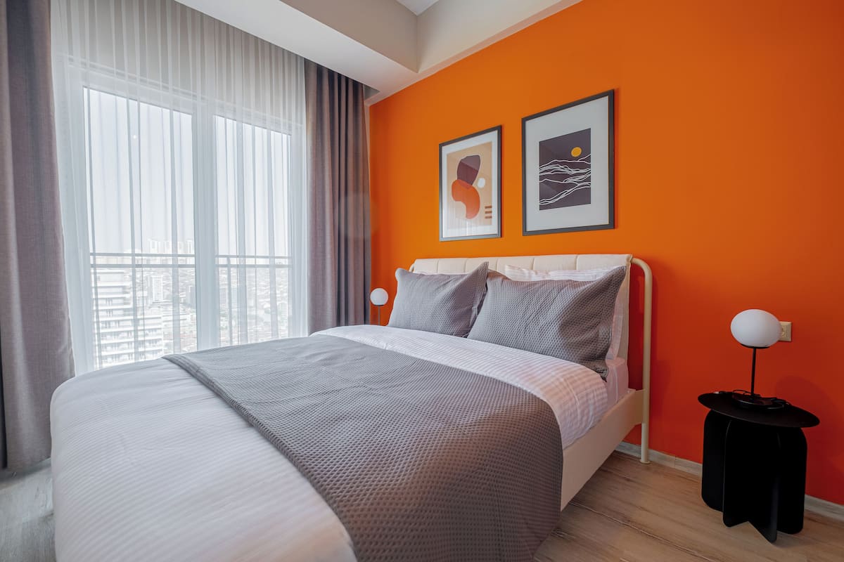 A modern hotel room with a large bed, a window with curtains, and two framed artworks on the orange wall.