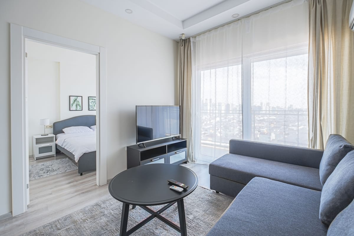 A modern hotel room with a bed, a TV, a sofa, and a view of the city through large windows.