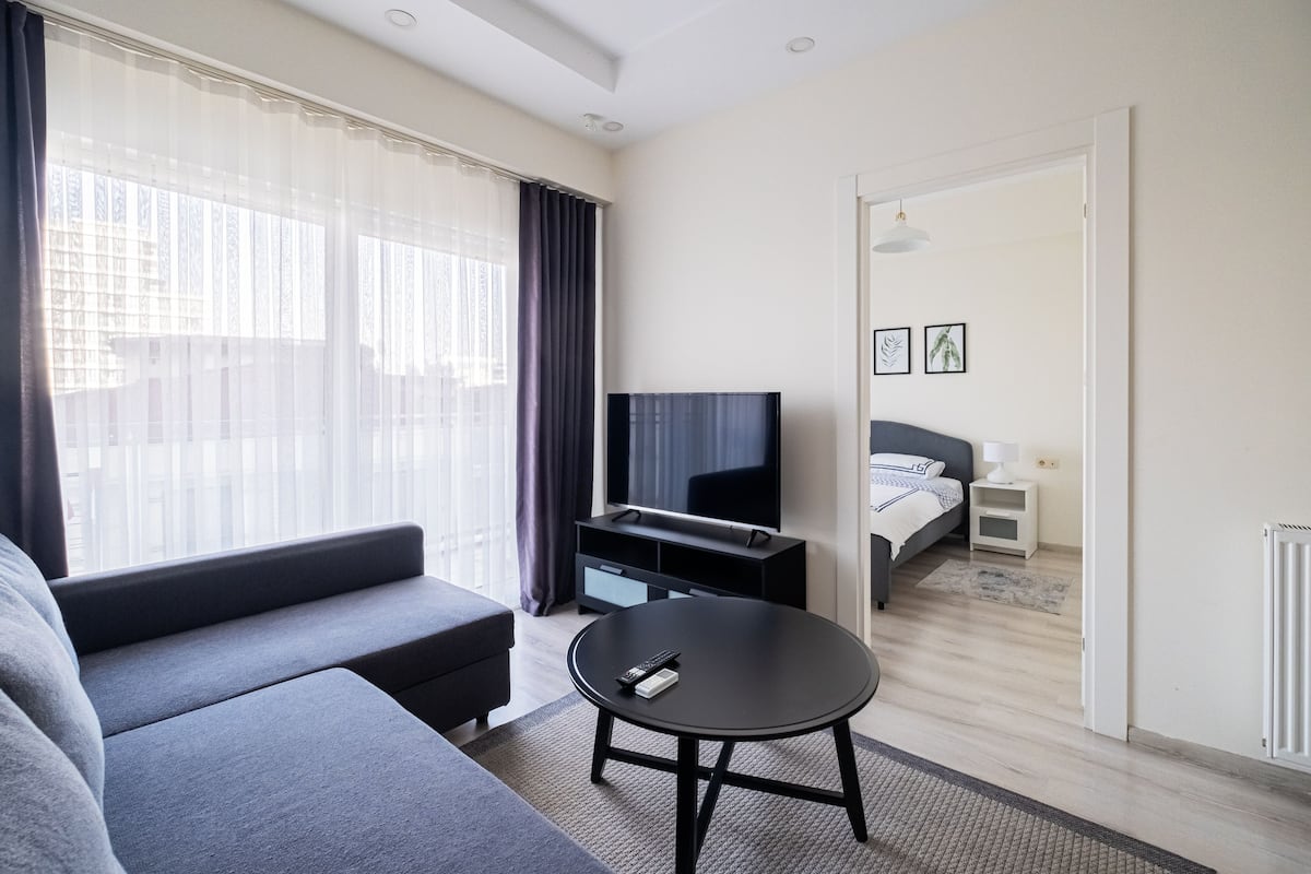 A modern hotel room with a sofa, a round black table, a TV, and a bed with a nightstand.