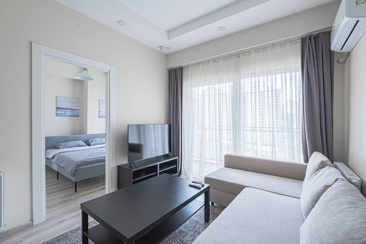 A modern hotel room with a bed, a TV, a sofa, and a large window with curtains.