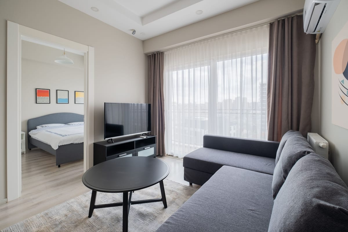 A modern hotel room with a bed, a TV, a sofa, and a dining table.