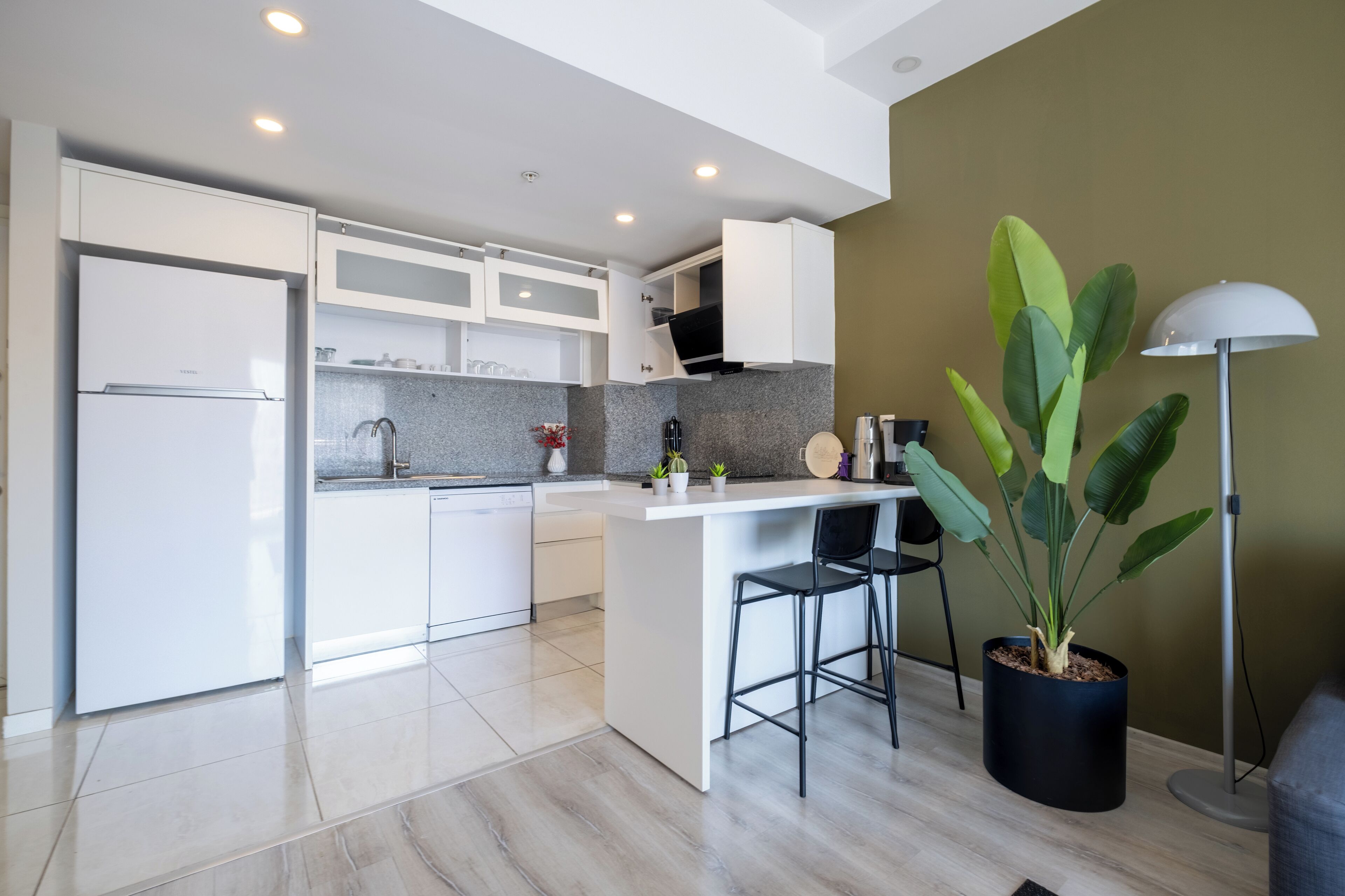 A modern kitchen with white cabinetry, a built-in island with bar stools, and a potted plant.