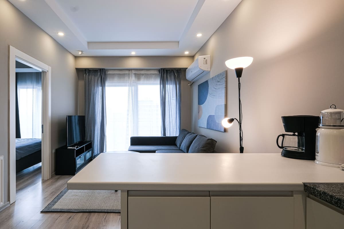 A modern hotel room with a large window, a flat-screen TV, a sofa, a coffee maker, and a lamp.