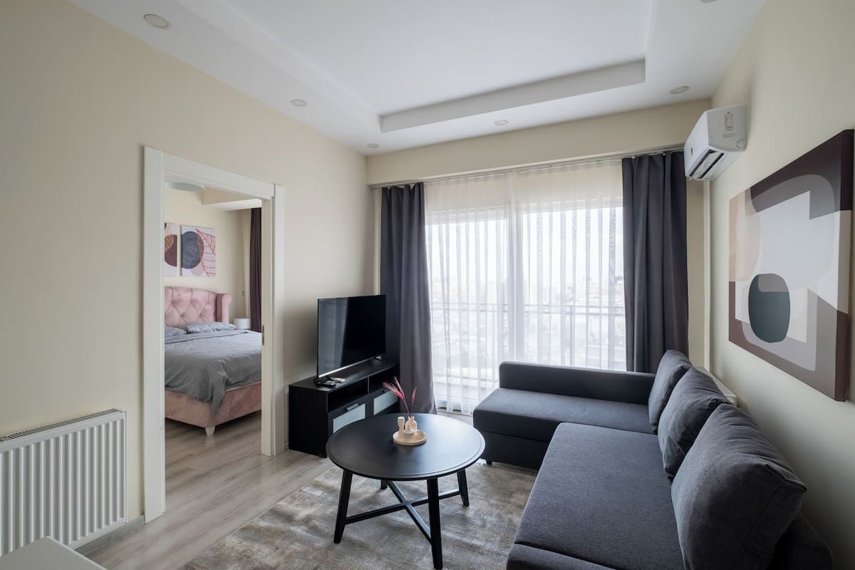 A modern living room with a sofa, a television, a small table, and a bed in the background.