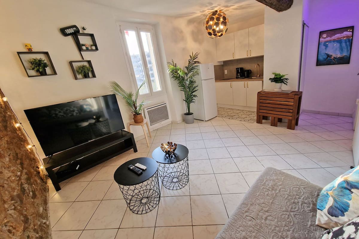 A living room with a TV, two wireframe tables, a sofa, and a kitchen area with cabinets and a hanging light fixture.