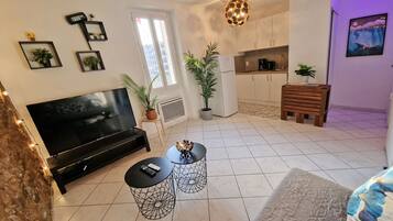 A living room with a TV, two wireframe tables, a sofa, and a kitchen area with cabinets and a hanging light fixture.