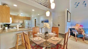 A modern kitchen with wooden cabinets, a central island, and a dining area with a glass table and chairs. - Island-Inspired 2bd with loft, lanais and serenity (Kihei)