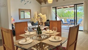 A dining area with a glass table, wooden chairs, and a view of the outdoor balcony. - Island-Inspired 2bd with loft, lanais and serenity (Kihei)