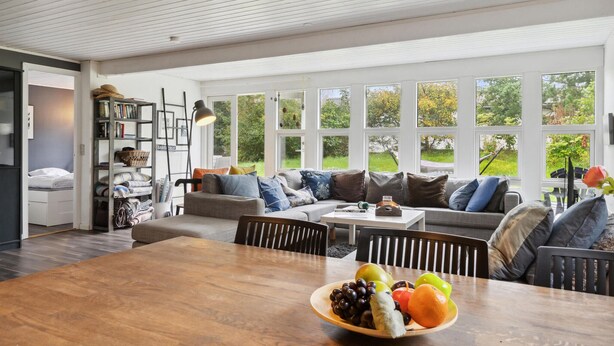 A spacious living room with a large wooden dining table, a grey sofa with multiple cushions, and a bookshelf filled with books and decorative items. - 4 bedroom awesome home in Sæby (Sæby)