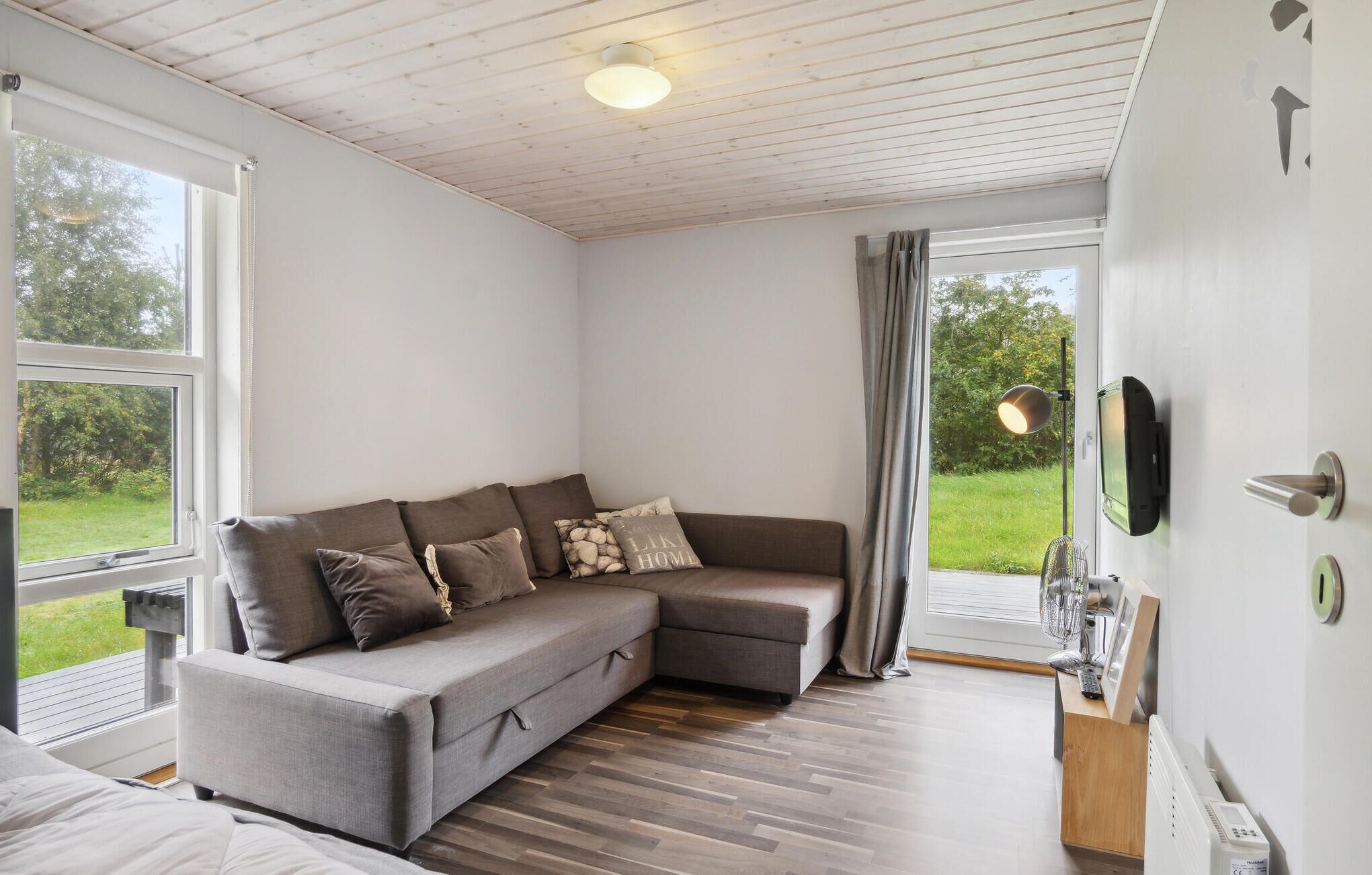 A modern living room with a grey sectional sofa, a flat-screen TV mounted on the wall, and a large window offering a view of the outdoors.