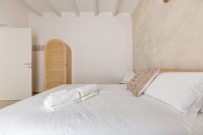 A neatly made bed with white linens and a rolled towel, a white door, and a woven wall panel.
