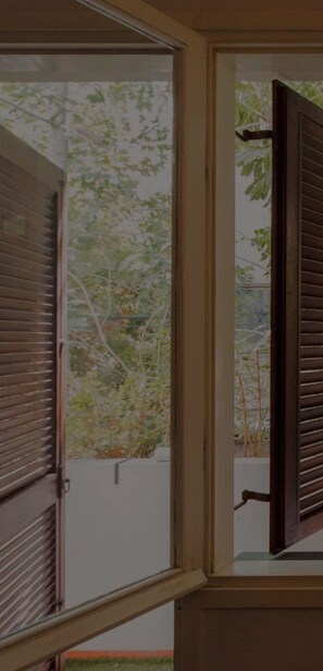 A window with wooden shutters partially open, revealing a view of a tree and a white wall outside.