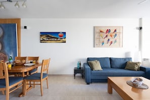 A living room with a blue sofa, wooden dining table, and chairs, a coffee table, and artwork on the walls.