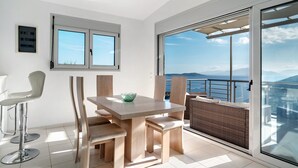 Ein Essbereich mit Tisch und Stühlen, ein Barhocker, ein Fenster mit Jalousien und Blick aufs Meer.