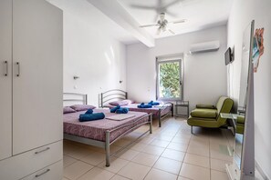 Une chambre avec deux lits, un fauteuil vert, un ventilateur de plafond et une fenêtre donnant sur des arbres.