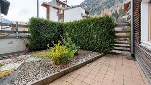 A modern building with a balcony, a garden with shrubs and a small plant, and a mountainous backdrop.
