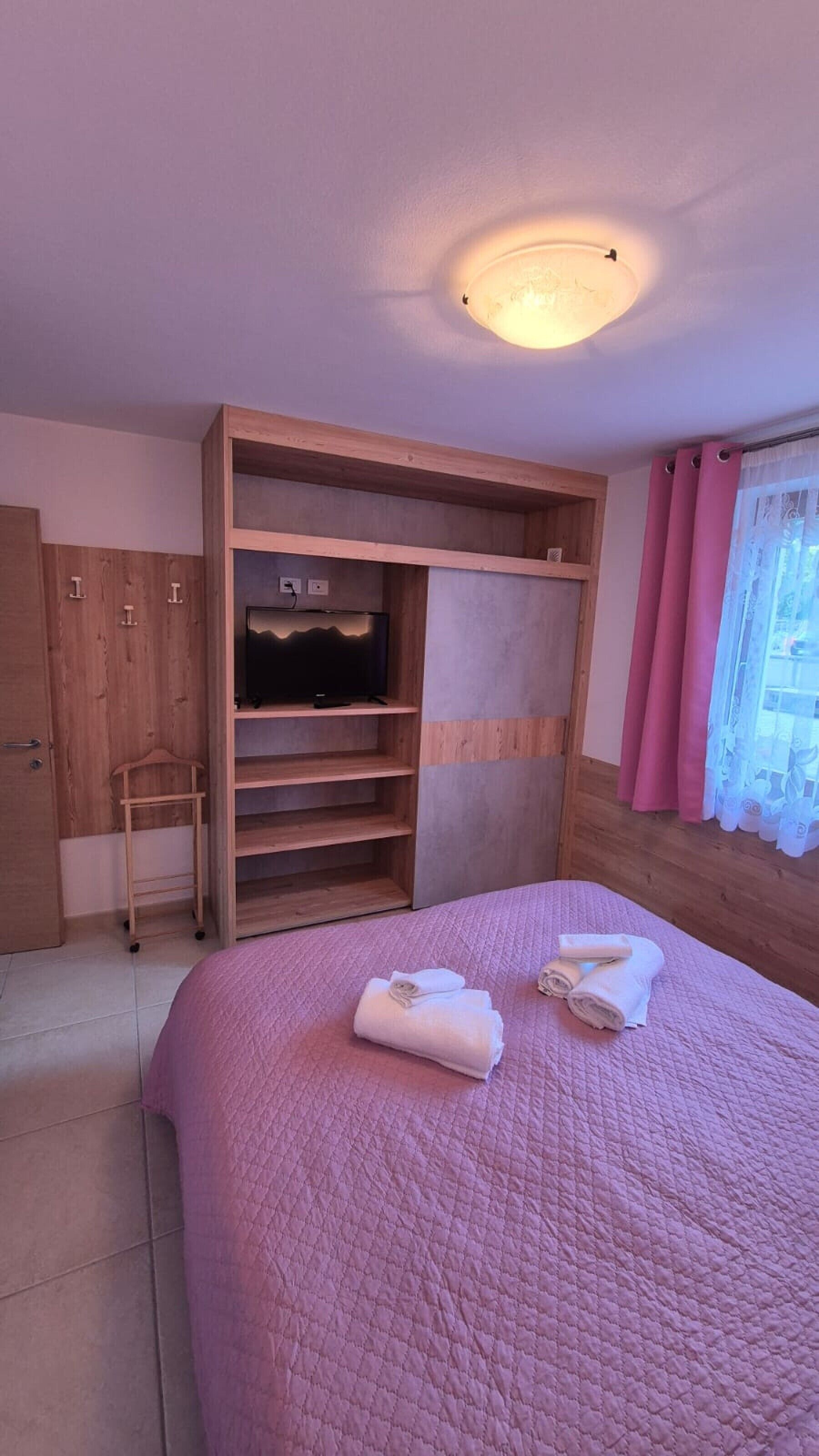 Ein Schlafzimmer mit einem Bett, einem Fernseher auf einem Holzboden, einem Kleiderschrank und einem Fenster mit Vorhängen.