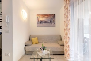 Een woonkamer met een bank, een glazen salontafel met een bloevas, een schilderij van een besneeerd landschap, en een telefoon aan de muur.