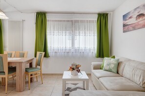 A living room with a dining table, chairs, a sofa, a coffee table with a flower vase, and a window with green curtains. - Apartment 'Night and Day 8' with Mountain View, Balcony and Wi-Fi (Predazzo)