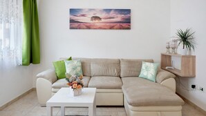 A living room with a beige sectional sofa, a white coffee table, and a painting of a sunset landscape. - Apartment 'Night and Day 8' with Mountain View, Balcony and Wi-Fi (Predazzo)