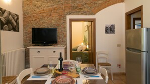 Een eethoek met een stenen muur, een televisie, een koelkast, en een tafel gedekt voor twee personen, inclusief borden, glazen en een fles.
