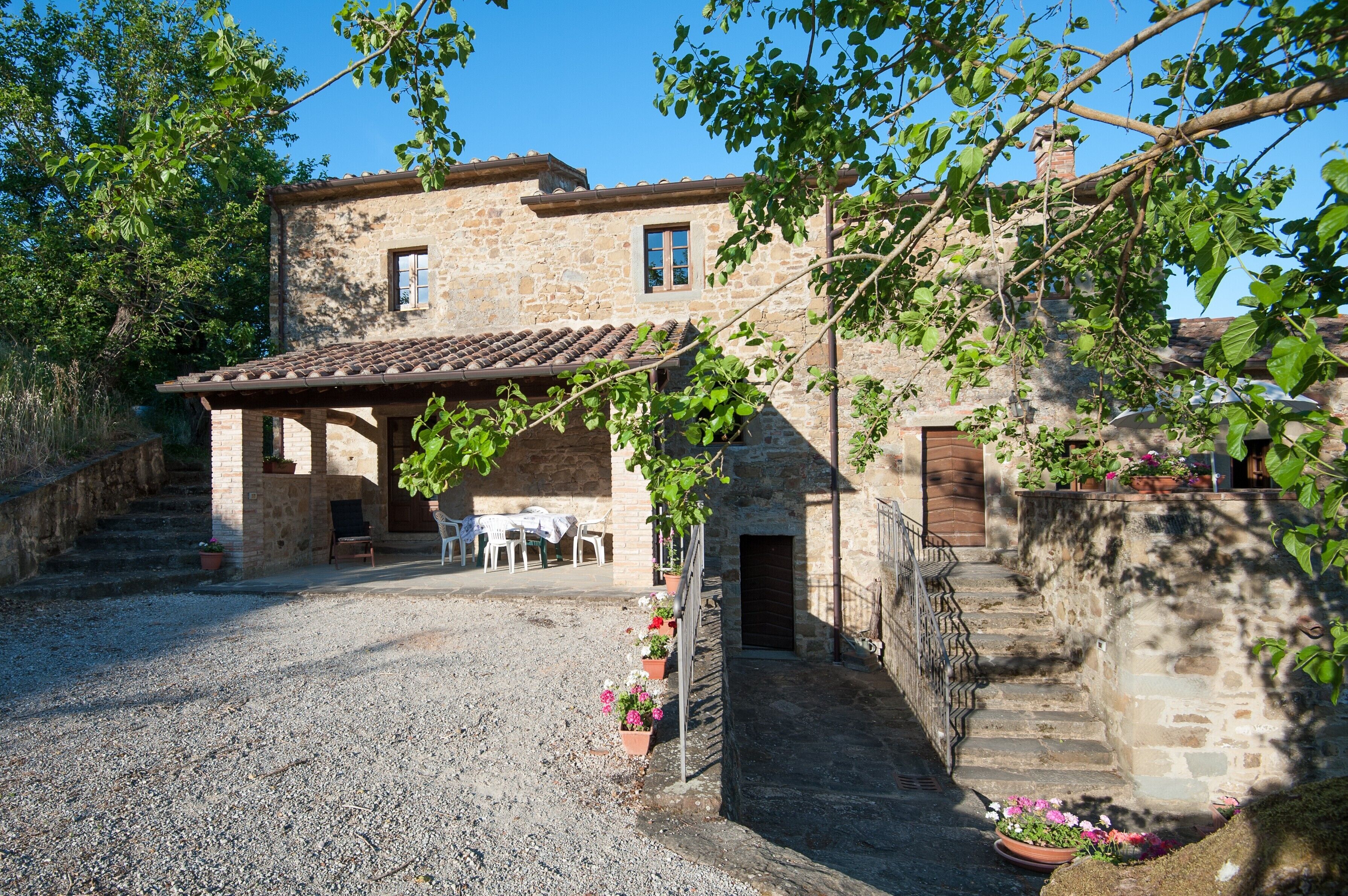 Un bâtiment en pierre avec un toit en tuiles, une allée gravillonnée et un petit espace extérieur aménagé avec des tables et des chaises.