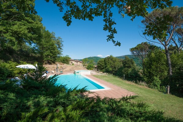 A swimming pool surrounded by lush greenery, a small building in the background, and a clear blue sky. - Farmhouse 'Pilari Appartamento Tabaccaia' with Mountain View, Shared Pool and Wi-Fi (Cortona)